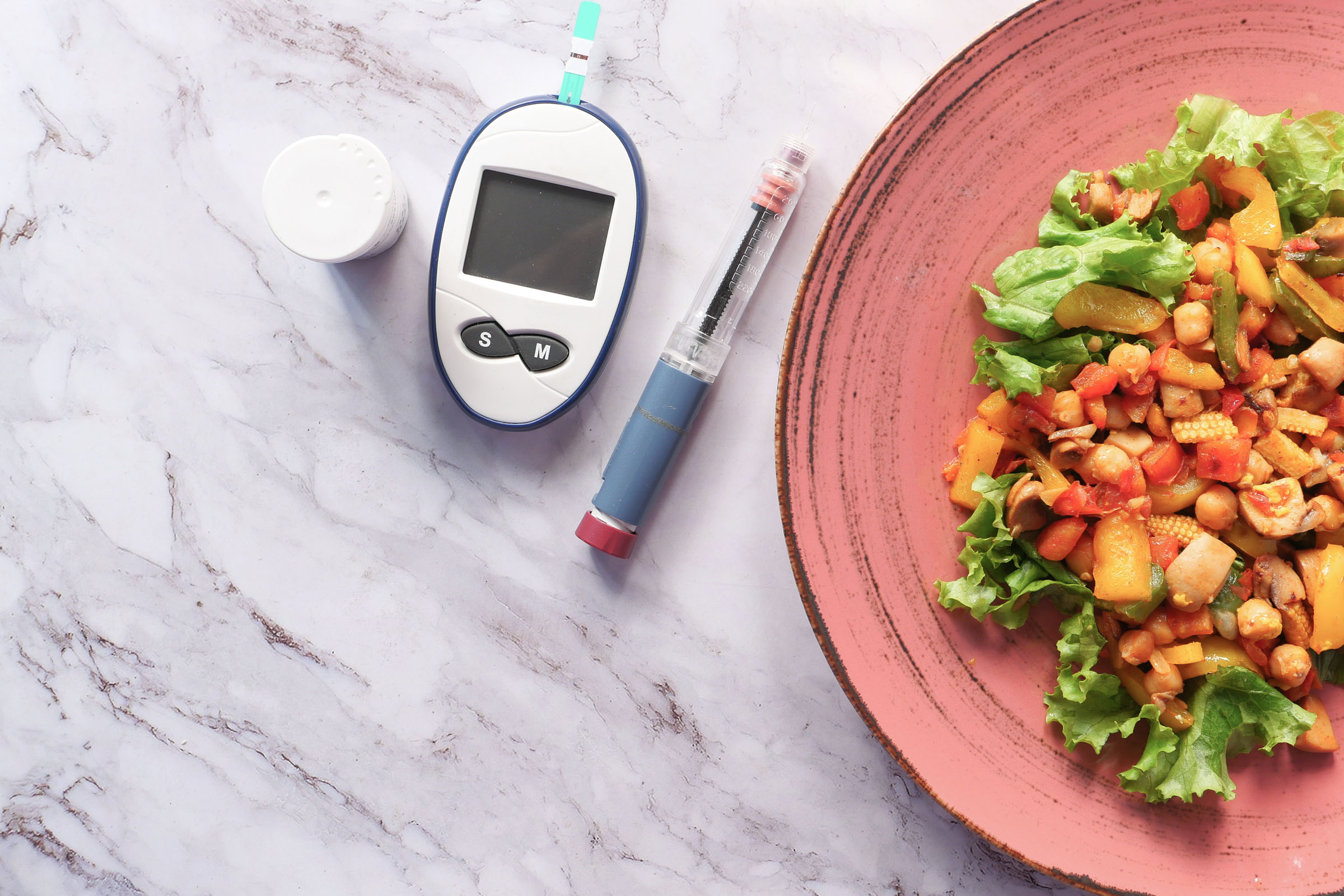 A glucose meter and insulin pen lie on a marble surface next to a colorful plate of healthy mixed vegetables, indicating balanced nutrition and diabetes management