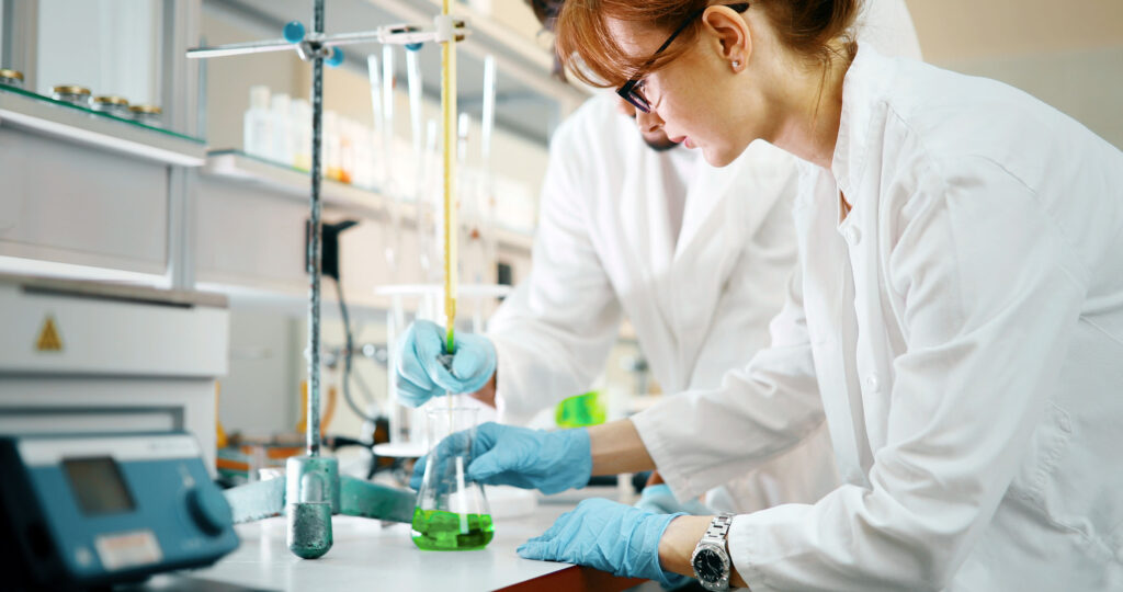 Two scientists in lab coats and blue gloves conducting an experiment with green liquid in a flask using laboratory glassware, working closely on a chemical procedure in a modern lab.
