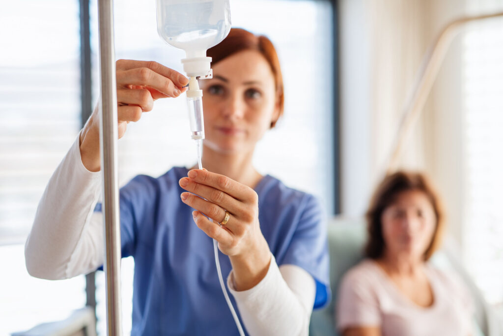 Nurse adjusting an IV drip in a clinical setting, with a seated patient blurred in the background, illustrating intravenous drug administration and hospital care.