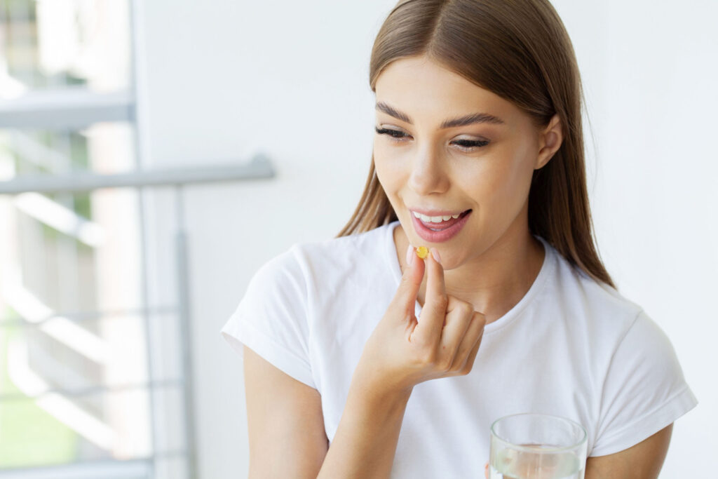 Smiling young woman in a white shirt holding a yellow capsule near her mouth with one hand and a glass of water in the other, preparing to take a supplement.