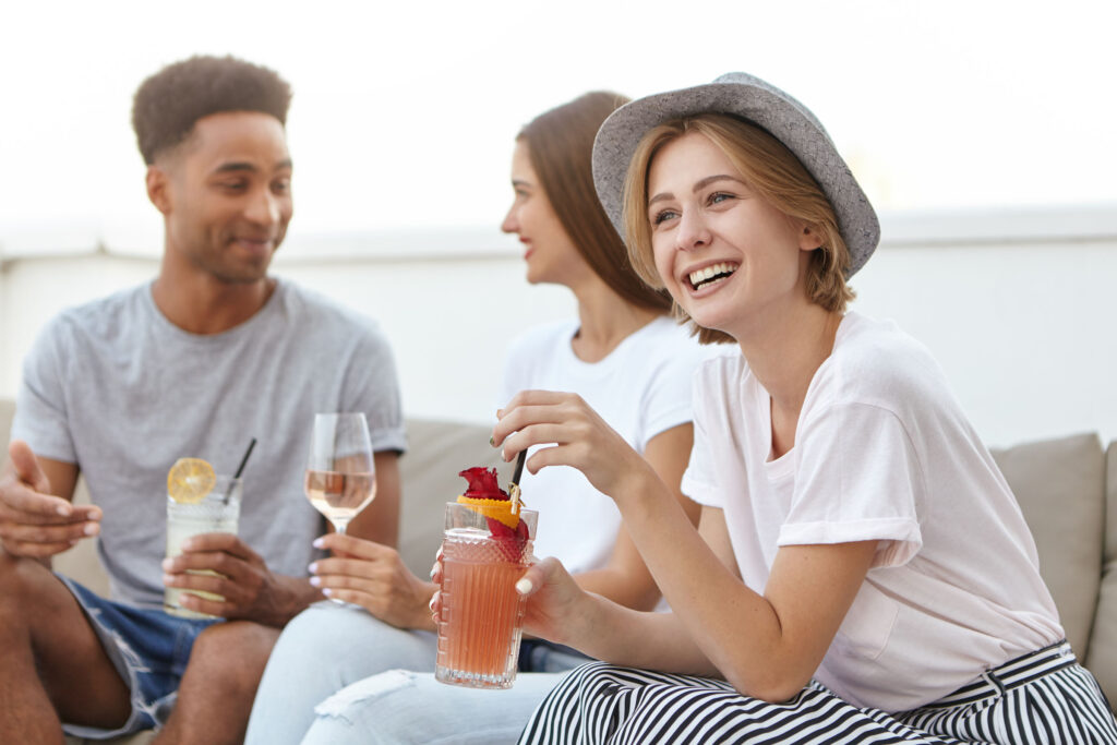 Group of young adults socializing and laughing outdoors while holding cocktails and wine glasses, creating a relaxed and joyful atmosphere.