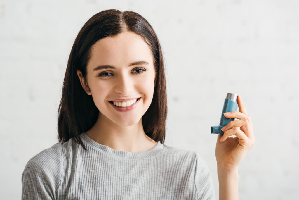 Smiling young woman holding a teal-colored asthma inhaler, standing against a softly lit background, representing respiratory health and confidence.