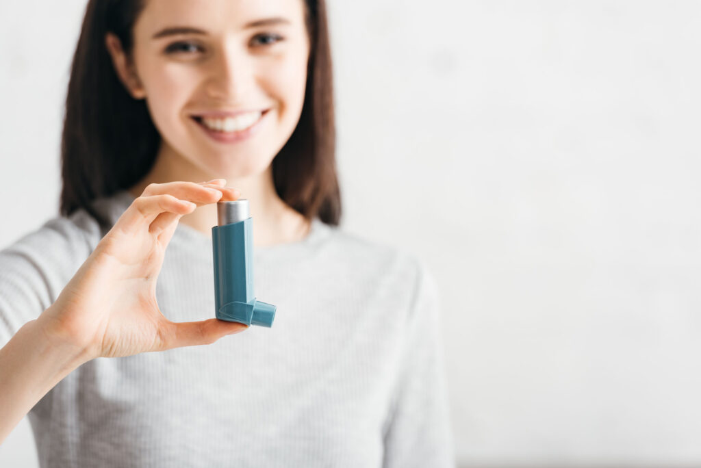 Close-up of a smiling woman holding a teal inhaler in focus, highlighting her hand and the device, with a blurred background suggesting healthcare support.
