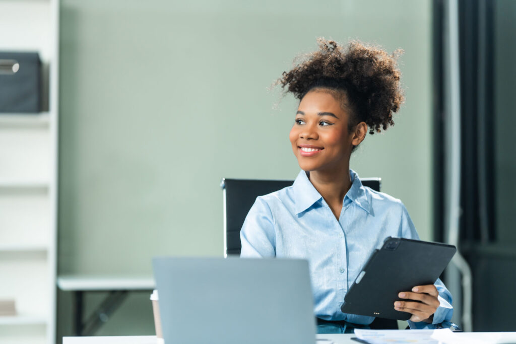 Smiling young professional woman in a light blue blouse holding a tablet, seated at a desk with a laptop and documents, looking off-camera in a modern office.