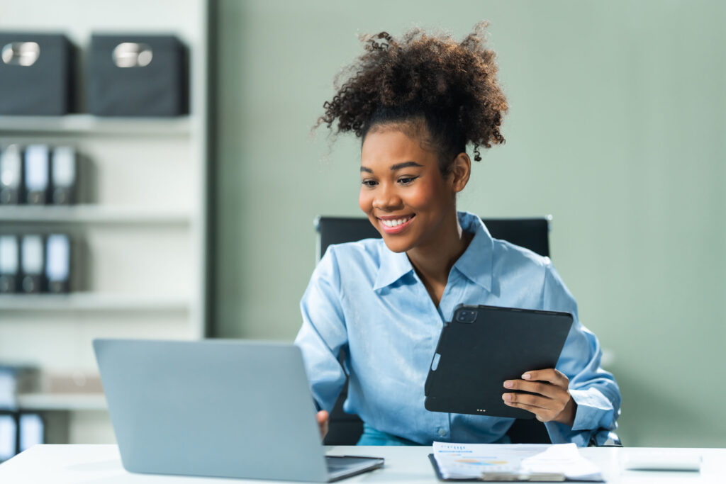 Smiling young professional woman reviewing content on a laptop with a tablet in hand, suggesting multitasking or virtual collaboration in a clean office environment.
