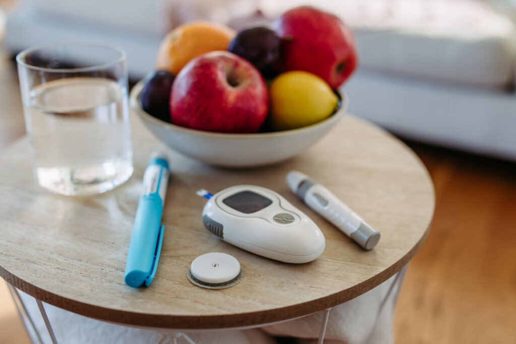 A tabletop with diabetes management tools including a glucometer, insulin pen, lancing device, and a continuous glucose monitor patch, next to a glass of water and a bowl of fresh fruit.