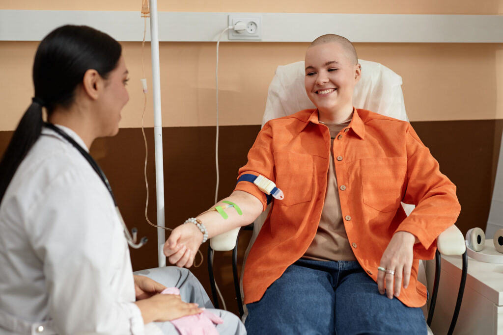 A smiling patient in an orange shirt receiving IV therapy in a clinic, seated and interacting with a medical professional in a white coat.