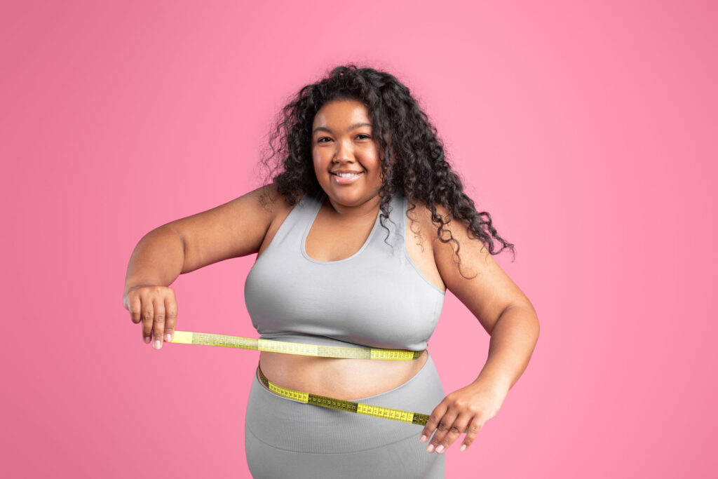Confident woman in activewear smiling while measuring her waist with a yellow measuring tape, standing in front of a pink background.